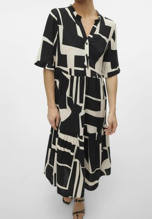 Frau trägt ein midi Kleid mit schwarz-beigem geometrischem Muster, Knöpfen auf der Brust und kurzen Rüschärmeln.