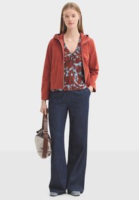 Veste à capuche rouge, blouse fleurie bleue et rouge avec un détail à nouer, pantalon large en denim foncé, et chaussures grises. Sac gris avec sangle marron.