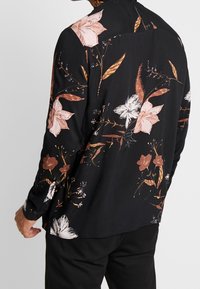 Chemise noire à manches longues avec un motif floral dans des teintes de rose, blanc et marron. Fabriquée en tissu léger avec une coupe décontractée.