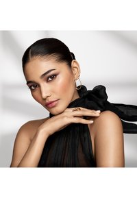 Lähikuva henkilöstä, jolla on päällään musta laskostettu hihaton paita. Koristeena kultaiset renkaat, ja sormus. Ihon pinta on sileä; tummat hiukset on sidottu taakse.