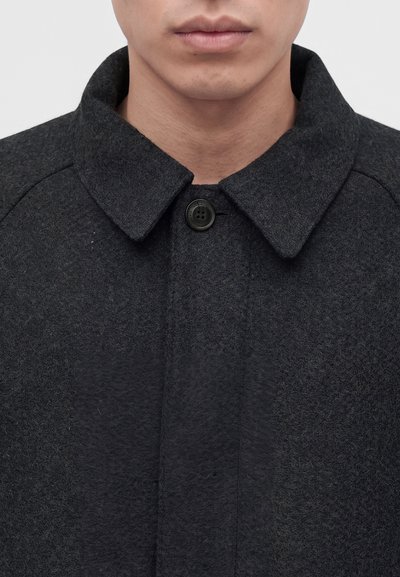 Man wearing a dark gray buttoned wool coat with a wide collar, showing part of the neck and lips against a plain light background.