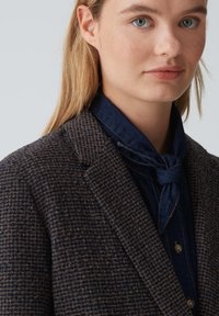 Blazer a motivo houndstooth marrone con revers, tessuto testurizzato e camicia di jeans blu scuro sotto, con dettagli di sciarpa annodata.