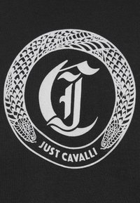 Ringikujuline madu muster ümbritseb stiliseeritud tähte "J", mille all on "JUST CAVALLI" tekst mustal kangasel taustal.