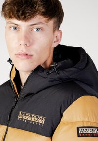 Giacca a vento nera e senape con chiusura a zip, cappuccio e logo ricamato; presenta una superficie liscia e texture, oltre a cuciture visibili.