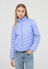 Giacca piumino viola chiaro con colletto alto, caratterizzata da un motivo a zigzag quilté e una cerniera frontale. Abbinata a jeans in denim blu.