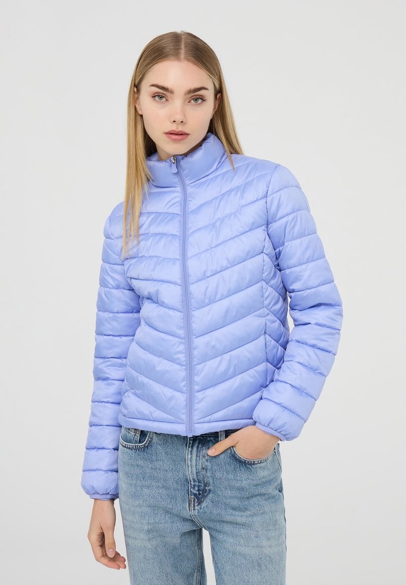 Giacca piumino viola chiaro con colletto alto, caratterizzata da un motivo a zigzag quilté e una cerniera frontale. Abbinata a jeans in denim blu.