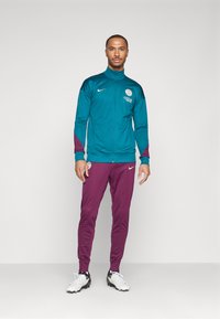 Teal zip-up takki maroon-yksityiskohtien ja logon kanssa, yhdistettyinä marooniin urheiluhousuihin. Malli käyttää valkoisia urheilukenkiä, joissa on mustia yksityiskohtia.