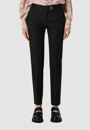 Donna che indossa pantaloni neri su misura con piega frontale, abbinati a mocassini neri con dettaglio a catena dorata e una camicetta rosa a fantasia.