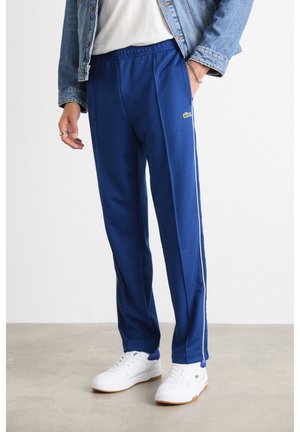 Hombre que lleva pantalones deportivos azules con rayas blancas a los lados, zapatillas blancas, una camiseta blanca y una chaqueta vaquera, de pie sobre un suelo gris.