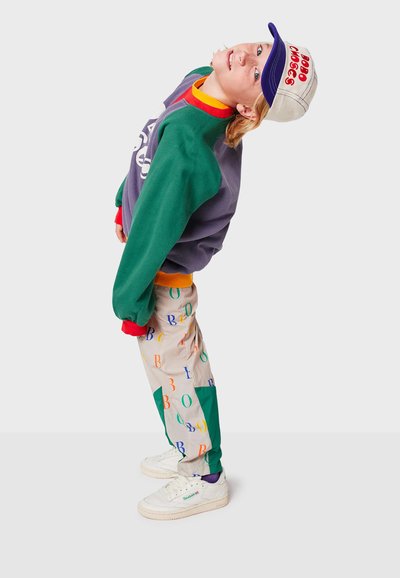 Outfit colorato composto da una felpa grigia con maniche verdi, pantaloni multicolori con lettere e sneakers bianche con dettagli viola.