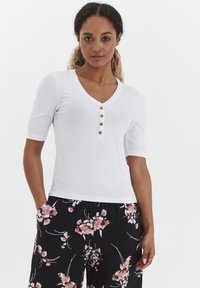 Hvid ribbet kortærmet top med V-hals og fem brune knapdetaljer, parret med sorte bukser med pink blomsterprint.