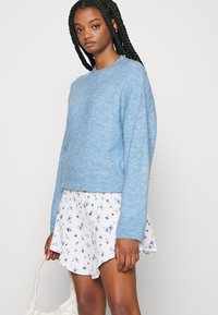 Suéter azul claro de malha com mangas longas e decote redondo, combinado com uma saia branca com padrão floral. Tecido texturado e corte relaxado.