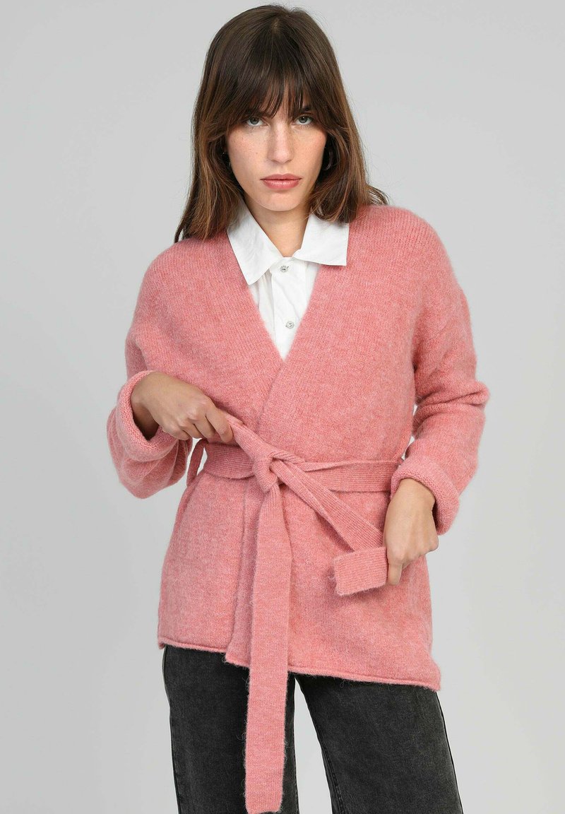 Gilet en tricot rose doux avec un design enveloppant, des manches longues et une ceinture assortie. Caractérisé par une texture douillette et un décolleté en V.
