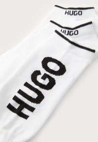Hvide bomuldsokker med et sort "HUGO" logo trykt fremtrædende på siden, med kontrasterende sorte striber øverst.