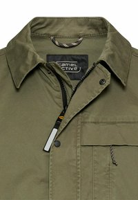 Olivengroene jas met kraag, gedeeltelijk open frontrits, drukknopen en een klepzak met trekkoord op de borst.