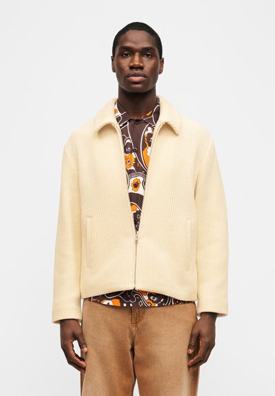 Άνδρας που φοράει μπεζ πλεκτό μπουφάν με φερμουάρ πάνω από καφέ, πορτοκαλί και λευκό πουκάμισο με λουλούδια, και ανοιχτό καφέ παντελόνι, στέκεται μπροστά σε απλό λευκό φόντο.