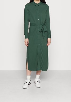 Femme portant une robe-chemise verte à manches longues boutonnée avec une ceinture à la taille, associée à des chaussettes blanches et des baskets blanches Adidas à rayures noires.