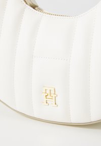 Vit läderhandväska med vertikala ränder, med en detaljerad logotyp i guldton och dragkedjestängning. Slät textur med en strukturerad form.