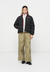 Chaqueta de plumas negra con detalles rojos y cuello con cremallera, combinada con pantalones cargo color beige que cuentan con grandes bolsillos y cremalleras.