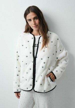 Femme portant une veste polaire blanche avec des motifs d'étoiles noires, bordure noire, fermeture éclair noire et un pantalon blanc à petits pois noirs.