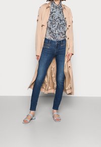 Trench beige sobre una blusa azul con volantes, combinada con jeans ajustados azul oscuro y sandalias azul claro. Tejido suave y corte entallado.
