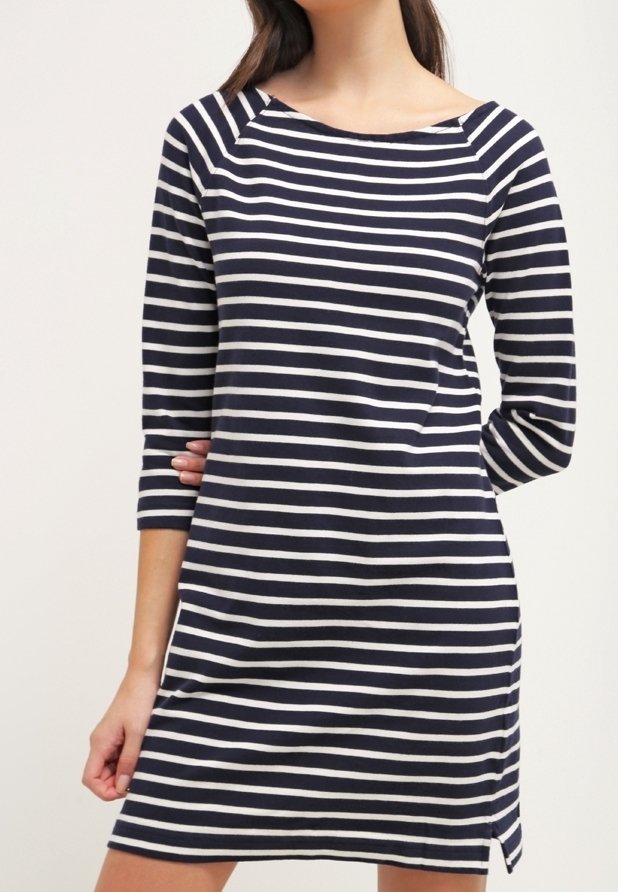 Femme portant une robe à rayures horizontales bleu marine et blanches, manches trois-quarts, avec une petite fente latérale, debout, une main derrière le dos.