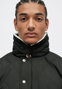 Giacca nera trapuntata con collare alto, bottoni a pressione argento e morbida fodera bianca intorno al colletto. Design semplice e funzionale.