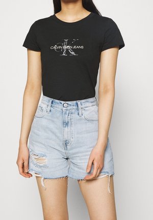 Kvinde iført en sort Calvin Klein Jeans t-shirt og lyseblå højttaljede distressed denimshorts, stående mod en hvid baggrund.