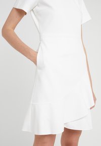 Femme portant une robe blanche à manches courtes avec un ourlet à volants superposés et la main dans la poche latérale, montrée du milieu du torse jusqu'aux genoux.