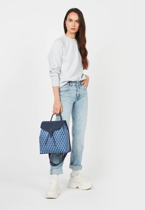 Sac à dos à motif bleu avec un rabat bleu foncé, en tissu texturé et une poignée supérieure, associé à un jean délavé et des baskets blanches.