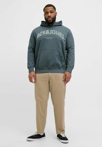 Mørk teal hættebluse med "ACK&JONES" tekst, frontlomme, parret med beige chinos og sorte canvas sneakers.