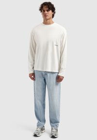 Witte langemouwen shirt met "THE PATH" in het zwart gedrukt op de linkerborst, gecombineerd met lichtblauwe relaxed-fit jeans en zilveren sneakers.