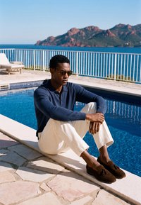 Homme portant des lunettes de soleil, une chemise bleu marine et un pantalon blanc, assis près d'une piscine avec des montagnes et une mer bleue en arrière-plan par une journée ensoleillée.
