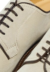 Scarpe in suede beige con lacci e dettagli in marrone scuro. Presentano una rifinitura in pelle liscia lungo i bordi e cuciture elaborate sui lati.
