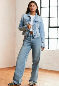 Chaqueta de mezclilla azul clara con cierre de botones, combinada con pantalones vaqueros de tiro alto en azul claro. Unos zapatos planos de rayas blancas y negras completan el atuendo.
