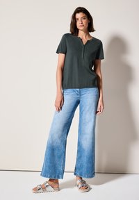 Haut vert foncé à manches courtes avec un col en V, associé à un jean large bleu clair. Le modèle porte des sandales argentées avec une sangle texturée.