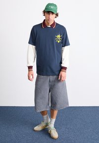 Marineblå polo med en bordeauxfarvet krave, der har et gult grafisk element, lagd over en hvid langærmet trøje. Parret med grå shorts og beige sneakers.