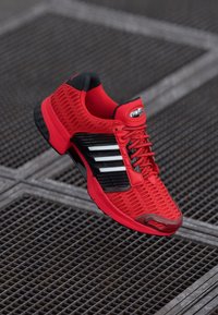 Red athletic shoe with mesh upper, black accents, and three white stripes. Features a textured sole and lacing system for a secure fit.