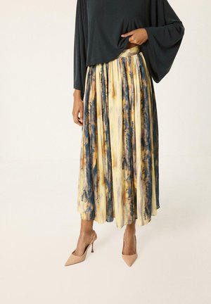 Femme portant une jupe midi plissée multicolore dans des tons jaunes, bleus et oranges, avec des talons hauts beiges et un haut à manches longues noir.