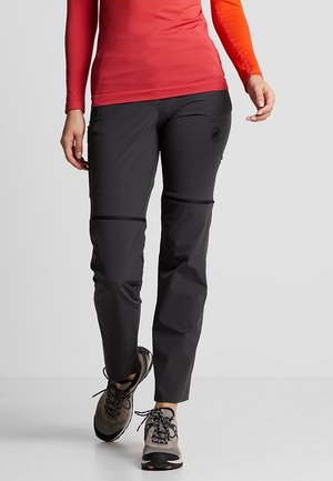 Person iført sorte vandrebukser med lynlås, orange langærmet skjorte og grå trail løbesko går fremad på en ensfarvet baggrund.