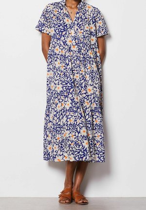 Frau trägt ein blaues Midikleid mit Blumenmuster, kurzen Ärmeln und braunen Sandalen mit Schnalle, steht vor einer weißen Wand.