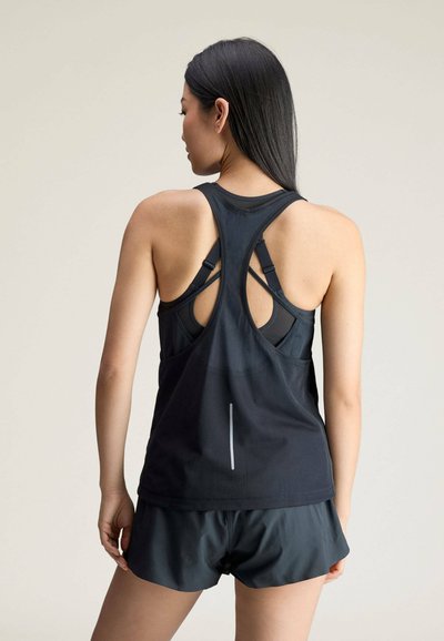 Woman with long dark hair wearing a black racerback tank top and black athletic shorts, standing with her back facing the viewer.