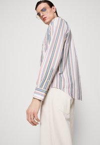 Camisa de manga larga a rayas en tonos blanco, rojo, azul y amarillo; presenta un cierre de botón en los puños y un dobladillo ligeramente curvado.