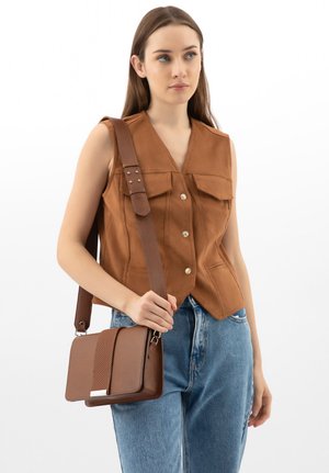 Kobieta nosząca bezrękawnik w kolorze brązowym z guzikami i niebieskie dżinsy, trzymająca brązową skórzaną torbę na ramię z szerokim paskiem.