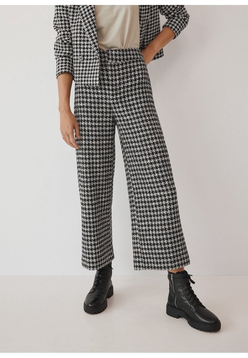 Femme portant un pantalon large à motif pied-de-poule noir et blanc, un blazer assorti, un haut beige et des bottines noires à lacets, debout devant un mur uni.