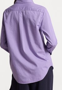 Camisa de manga larga color púrpura claro con cuello abotonado, presenta un pliegue central en la parte trasera y un dobladillo curvo. Hecha de una tela suave y texturizada.