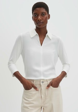 Personne portant une chemise blanche à manches longues avec col, rentrée dans un pantalon beige, mains dans les poches, debout devant un fond uni.