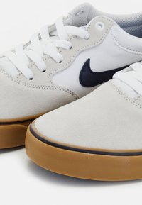 Baskets blanches et gris clair avec une tige en suède, des lacets blancs, un logo Swoosh bleu marine et une semelle extérieure en caoutchouc marron. Présentent un design à bout arrondi.