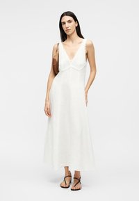 Nő ujjatlan fehér maxi ruhában, horgolt felsőrésszel, fekete pántos szandálban és barna válltáskával, egyszínű háttér előtt.