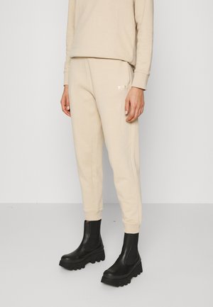 Persoon draagt beige sweatpants met een logo op de dij, een bijpassende beige sweater en zwarte grove plateau laarzen op een grijze vloer.